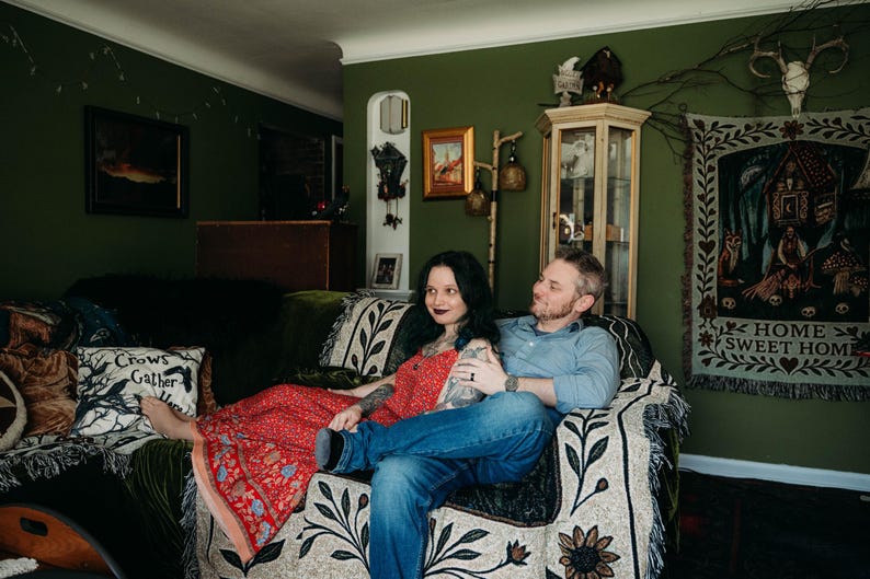 May include: A person in a red patterned maxi dress and a person in a blue button-down shirt and blue jeans sitting on a couch covered with a cream-colored throw blanket featuring a black floral print and a pillow with the text Crows Gather Here.