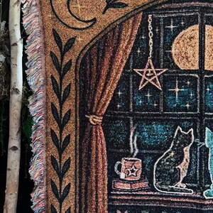 May include: Woven tapestry throw blanket featuring a celestial design with a crescent moon, stars, and a full moon. The blanket depicts a cat sitting by a window with a cup of coffee. The blanket has a fringe edge.