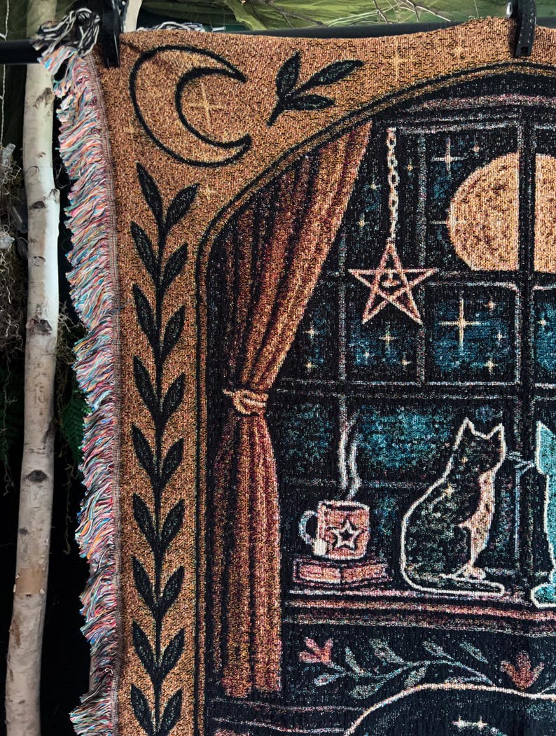 May include: Woven tapestry throw blanket featuring a celestial design with a crescent moon, stars, and a full moon. The blanket depicts a cat sitting by a window with a cup of coffee. The blanket has a fringe edge.
