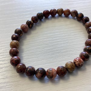 K&ouml;nnte beinhalten: Ein Armband aus runden, polierten Perlen in Braun-, Rot- und Beiget&ouml;nen. Das Armband ist aufgef&auml;delt und bildet eine kreisf&ouml;rmige Form. Die Perlen haben ein gesprenkeltes Aussehen.