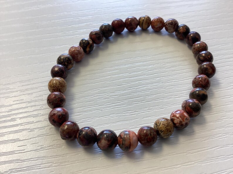 K&ouml;nnte beinhalten: Ein Armband aus runden, polierten Perlen in Braun-, Rot- und Beiget&ouml;nen. Das Armband ist aufgef&auml;delt und bildet eine kreisf&ouml;rmige Form. Die Perlen haben ein gesprenkeltes Aussehen.