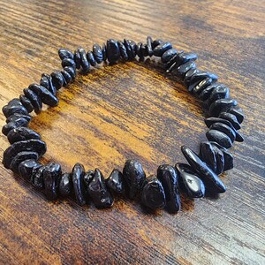 May include: A black gemstone bracelet composed of irregularly shaped, polished stones. The bracelet is strung together, forming a circular shape. The stones are a deep, glossy black, and the bracelet rests on a wooden surface.