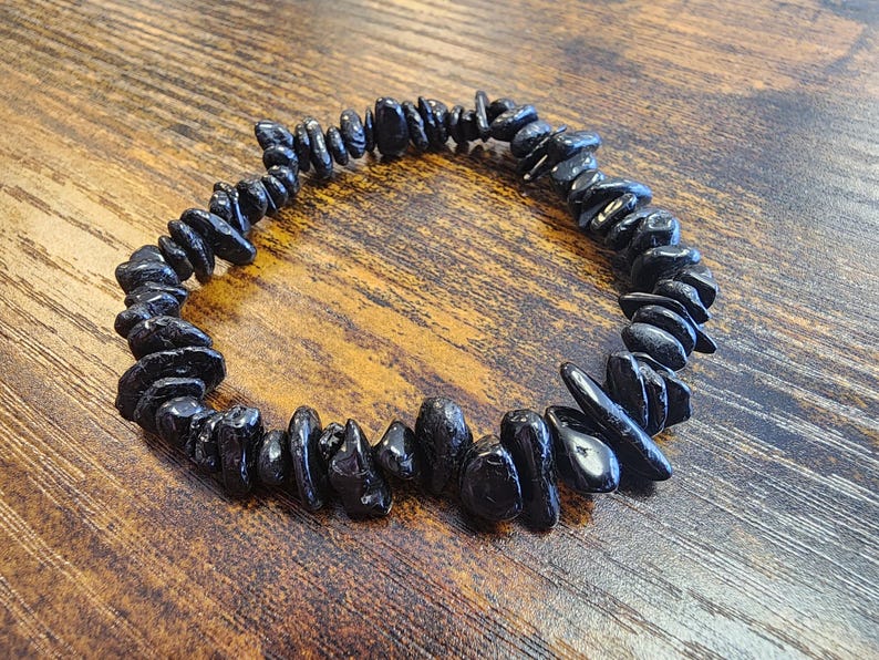 May include: A black gemstone bracelet composed of irregularly shaped, polished stones. The bracelet is strung together, forming a circular shape. The stones are a deep, glossy black, and the bracelet rests on a wooden surface.