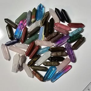 May include: A collection of approximately 40 small, pointed crystal wands in various colors, including pink, blue, green, purple, red, brown, and black. The wands are arranged in a random pile on a white surface.