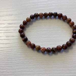 K&ouml;nnte beinhalten: Ein Armband aus runden, polierten Perlen in Braun-, Rot- und Beiget&ouml;nen. Die Perlen sind auf einem Gummiband aufgef&auml;delt. Das Armband liegt auf einer wei&szlig;en Oberfl&auml;che. Die Perlen haben ein gesprenkeltes Aussehen.