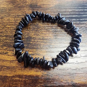 May include: A black gemstone bracelet composed of irregularly shaped, polished stones. The bracelet is strung together, forming a circular shape. The dark stones contrast against the wooden surface, creating a natural and earthy aesthetic.