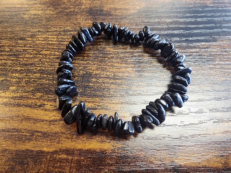 May include: A black gemstone bracelet composed of irregularly shaped, polished stones. The bracelet is strung together, forming a circular shape. The dark stones contrast against the wooden surface, creating a natural and earthy aesthetic.
