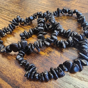 May include: Three black tourmaline chip bracelets are arranged on a wooden surface. The bracelets are made of small, irregularly shaped black stones strung together. The dark stones have a glossy finish and are strung on an elastic band.