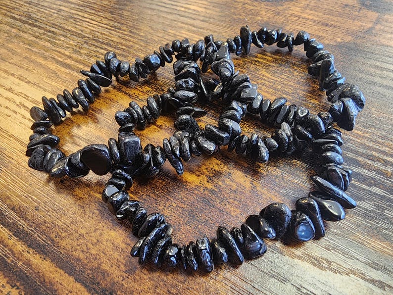 May include: Three black tourmaline chip bracelets are arranged on a wooden surface. The bracelets are made of small, irregularly shaped black stones strung together. The dark stones have a glossy finish and are strung on an elastic band.