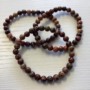 K&ouml;nnte beinhalten: Drei Armb&auml;nder mit Perlen in Braun-, Rot- und Beiget&ouml;nen. Die runden Perlen sind aufgef&auml;delt und bilden eine kreisf&ouml;rmige Form. Die Armb&auml;nder liegen auf einer wei&szlig;en Oberfl&auml;che.