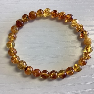 Puede incluir: Una pulsera de cuentas redondas de color ámbar. Las cuentas varían en tonalidad, desde amarillo claro hasta marrón oscuro, algunas con patrones internos. La pulsera está ensartada, creando una forma circular.