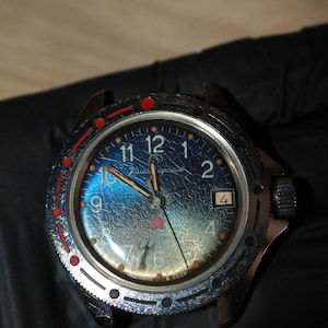 Puede incluir: Un reloj de pulsera vintage negro y plateado con una esfera azul y detalles rojos. La esfera del reloj tiene un efecto de cristal agrietado y presenta el texto "Чайка СССР".