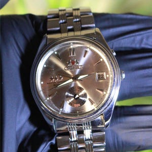 Puede incluir: Un reloj de pulsera plateado con una esfera plateada y agujas negras. El reloj tiene una ventana de fecha en la posición de las 3 en punto y una ventana del día de la semana en la posición de las 4 en punto. El reloj también tiene un pequeño subdial en la posición de las 6 en punto. El reloj está en una banda de metal plateado.