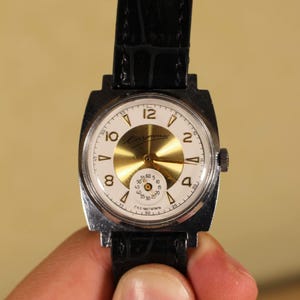 Puede incluir: Un reloj de pulsera vintage con esfera blanca, centro dorado y correa de cuero negro. El reloj tiene una caja rectangular plateada y muestra la hora con números negros. La esfera del reloj incluye el texto "Caymate".