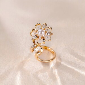 Puede incluir: Un anillo de tono dorado con un diseño en forma de flor, una perla blanca central y piedras claras y brillantes alrededor. El anillo es abierto y presenta detalles en forma de hoja adornados con pequeñas piedras claras a lo largo del lateral.