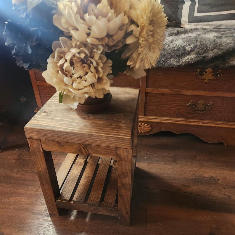 May include: A small, rustic wooden side table with a lower shelf. The table is stained a warm brown color and has a simple, boxy design. A vase of cream-colored flowers sits on top. The table is 18 inches tall.