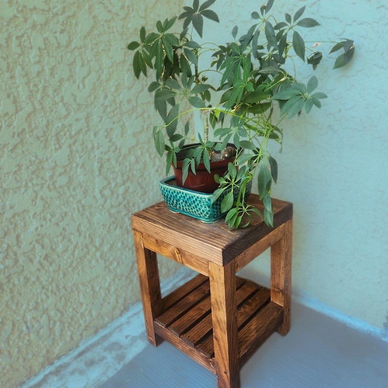May include: A small, rustic wooden plant stand with a lower shelf. A potted plant with green leaves sits on top, in a teal ceramic planter. The stand is made of stained wood and is placed against a textured wall.