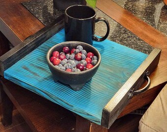 May include: A blue wooden tray with black trim and a metal handle. The tray is on a wooden table and has a black mug and a bowl of red berries and blueberries on it.