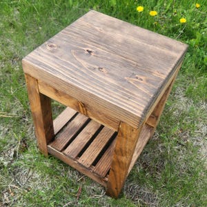 May include: A small, square wooden side table with a dark brown stain. The table has a slatted shelf and is constructed from thick planks of wood. The table is sitting on green grass.