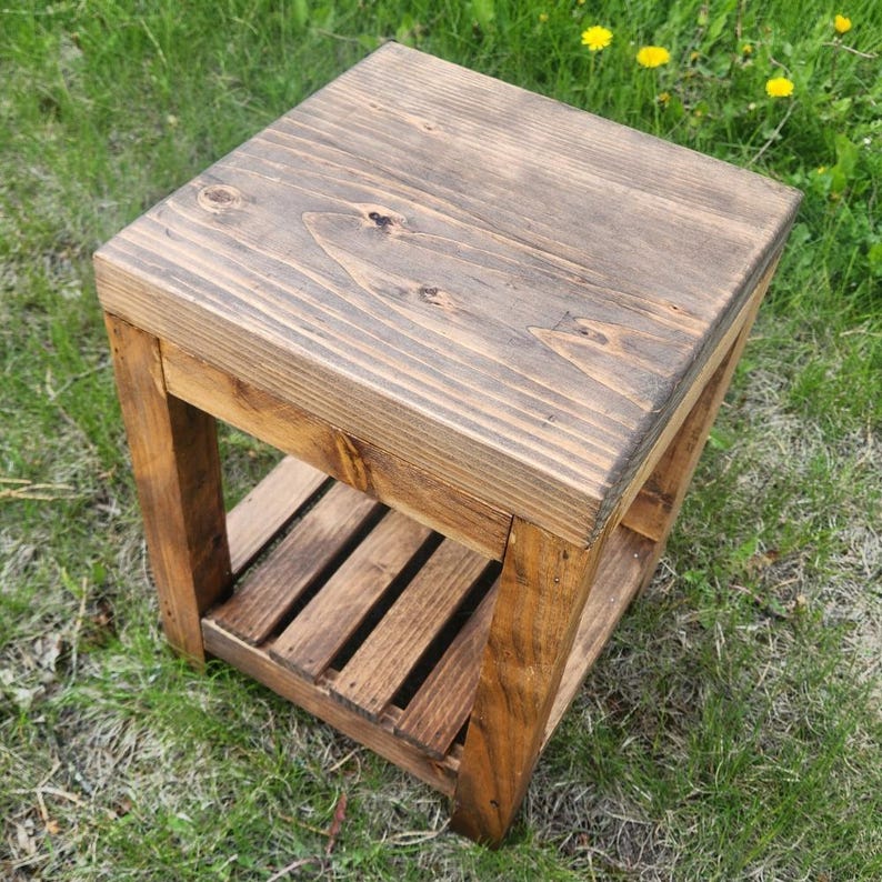 May include: A small, square wooden side table with a dark brown stain. The table has a slatted shelf and is constructed from thick planks of wood. The table is sitting on green grass.