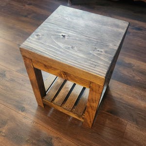 May include: A small, rustic wooden side table with a rectangular top and a slatted shelf. The table is stained in a warm brown color with a gray-toned top. The wood grain is visible, adding to the natural aesthetic. The table is designed for indoor use.