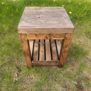 May include: A small, square wooden side table with a dark stain. The table has a slatted shelf and is constructed from planks of wood. The table is sitting on a bed of green grass.