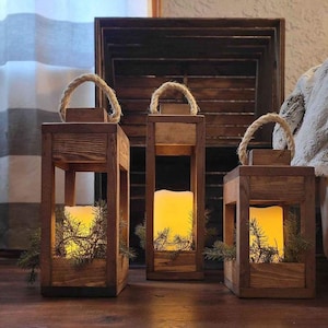 May include: Three wooden candle lanterns with rope handles and lit candles inside. The lanterns are made of rustic wood and have a natural finish. The candles are white and are surrounded by green pine branches.