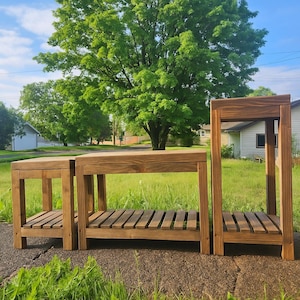May include: Three wooden plant stands of varying heights are displayed outdoors. The stands are constructed from stained wood, featuring a slatted shelf and a solid top. The tallest stand is approximately 36 inches high.