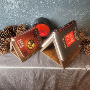May include: Two wooden bookends in the shape of a triangle, each holding a book. The book on the left is "Catching Fire" by Suzanne Collins. The book on the right is "The Art of War" by Sun Tzu.
