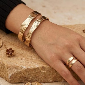 May include: Two gold-toned metal cuff bracelets with a hammered, textured surface worn on the wrist, paired with two matching gold-toned metal rings with a hammered finish worn on the finger. The jewelry pieces feature a wavy, organic edge design.