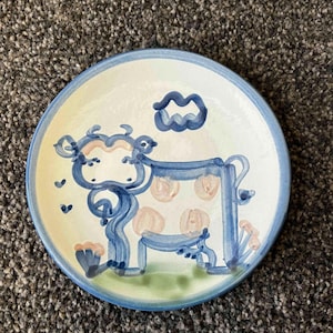 Puede incluir: Un plato de cerámica azul y blanco con un diseño pintado a mano de una vaca con manchas rosas. La vaca está de pie en un campo verde con un cielo azul arriba.