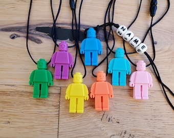 May include: Seven colorful silicone necklaces with a Lego-style figure pendant. Each necklace has a black cord and a different color figure: green, purple, blue, orange, yellow, pink, and light blue. One necklace has a name tag with the letters "NAME" in white.