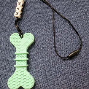 May include: A light green silicone teething toy shaped like a bone with a black cord and beads that spell "NAME".