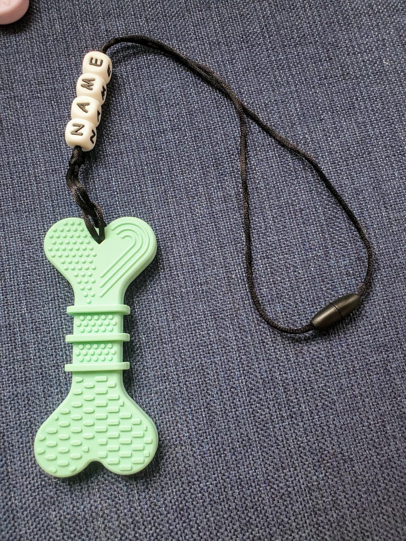 May include: A light green silicone teething toy shaped like a bone with a black cord and beads that spell "NAME".