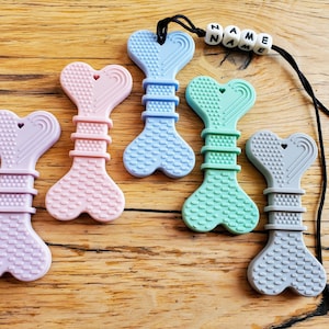 May include: Five silicone teethers shaped like bones with heart-shaped ends. The teethers are in various colors, including purple, pink, blue, green, and gray. The gray teether is attached to a black cord with white beads that spell "NAME".
