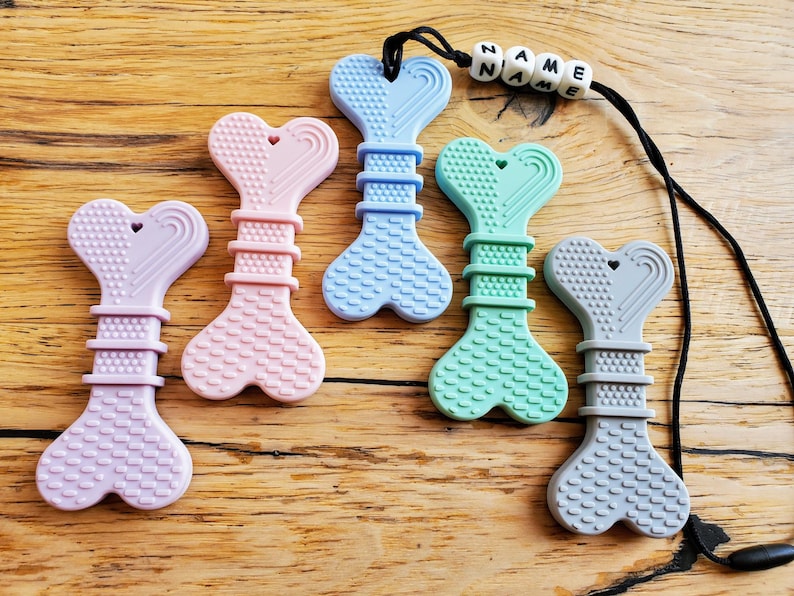 May include: Five silicone teethers shaped like bones with heart-shaped ends. The teethers are in various colors, including purple, pink, blue, green, and gray. The gray teether is attached to a black cord with white beads that spell "NAME".