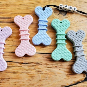 May include: Five silicone teethers shaped like dog bones in various colours: lavender, pink, light blue, mint green, and grey. Each teether has a heart-shaped design and a textured surface. The teethers are arranged on a wooden surface.
