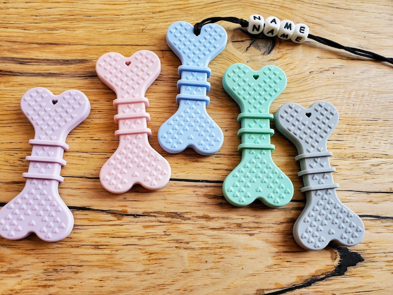 May include: Five silicone teethers shaped like dog bones in various colors: lavender, pink, light blue, mint green, and gray. Each teether has a heart-shaped design and a textured surface. The teethers are arranged on a wooden surface.
