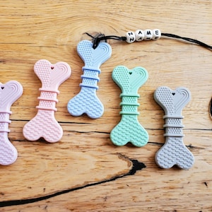 May include: Six silicone teething toys shaped like bones with a heart cutout. The toys are in various colors, including pink, blue, green, and gray. The toys are attached to a black cord with a name tag that reads "NAME".