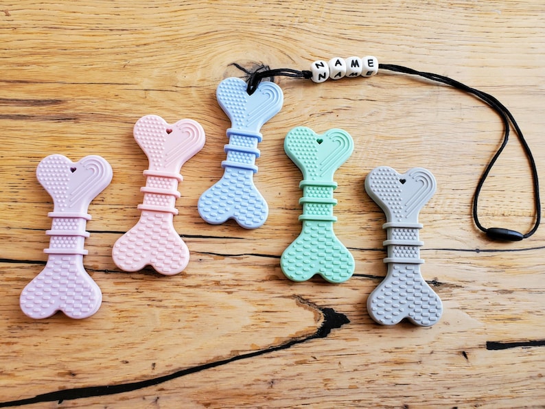 May include: Six silicone teething toys shaped like bones with a heart cutout. The toys are in various colors, including pink, blue, green, and gray. The toys are attached to a black cord with a name tag that reads "NAME".