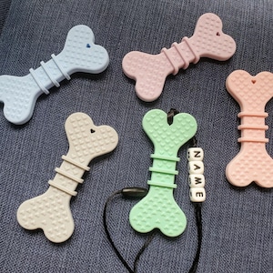 May include: Five silicone teethers shaped like bones in various colors, including light blue, beige, light green, and pink. Each teether has a heart-shaped design and is textured with small hearts. One teether has a black cord with a name bead that reads "NAME".