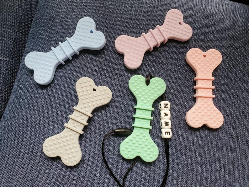 May include: Five silicone teethers shaped like bones in various colors, including light blue, beige, light green, and pink. Each teether has a heart-shaped design and is textured with small hearts. One teether has a black cord with a name bead that reads "NAME".