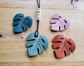 May include: Four silicone monstera leaf teethers in teal, pink, brown, and green. The teal leaf is attached to a black cord with white beads that spell "NAME".