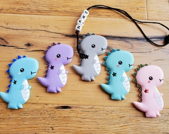 May include: Five colorful silicone dinosaur teethers with cute faces and star details. The teethers are attached to a black cord with a white bead necklace that spells "NAME".