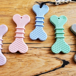 May include: Five silicone teething toys shaped like bones with a heart at the top. The toys are in the colors lavender, pink, light blue, green, and gray.