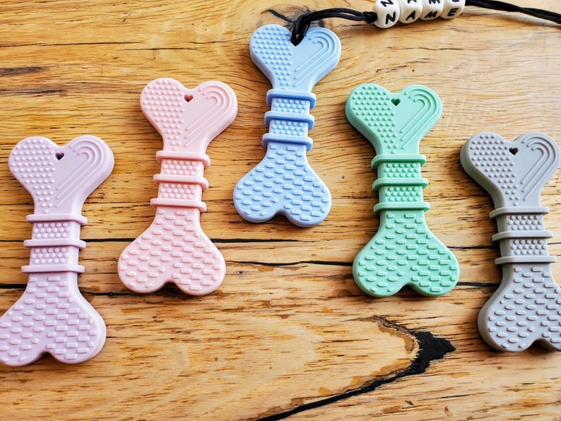 May include: Five silicone teething toys shaped like bones with a heart at the top. The toys are in the colors lavender, pink, light blue, green, and gray.