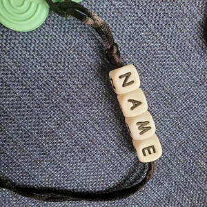 May include: A black cord with white beads that spell out the word "NAME".