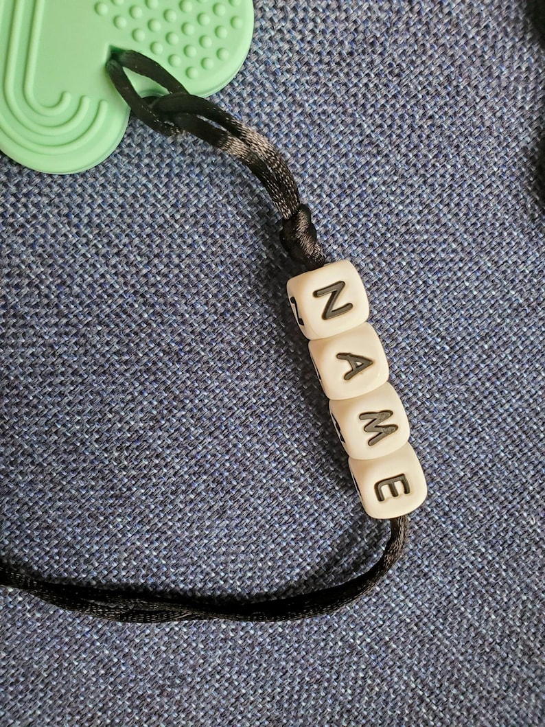 May include: A black cord with white beads that spell out the word "NAME".