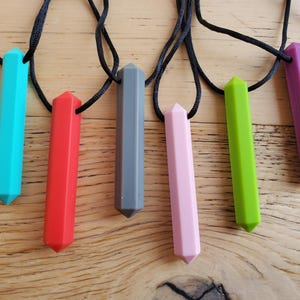 May include: Six silicone necklace pendants in various colours, including teal, red, grey, pink, green, and purple. Each pendant is shaped like a long, pointed hexagon.