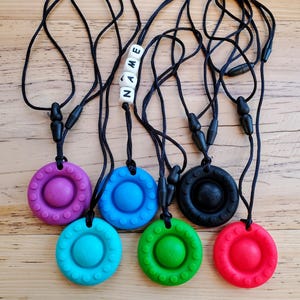 以下が含まれることがあります： 異なる色のペンダントが付いたシリコン製の噛みしめネックレスが6本。ペンダントは丸型で、中央が盛り上がっています。ネックレスは黒色で、黒いプラスチック製の留め具が付いています。ネックレスの1本には、「NAME」という文字が白いビーズで綴られています。